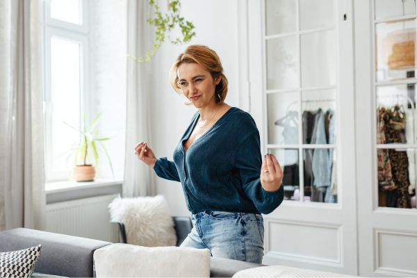 Frau bewegt sich locker und gut gelaunt im Wohnzimmer, macht eine kleine Tanz- oder Lockerungsbewegung neben einem Sofa.